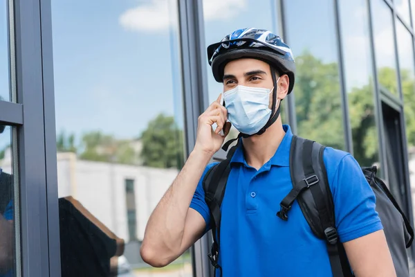 Arabo fattorino in casco da bici e maschera medica che parla su smartphone su strada urbana — Foto stock