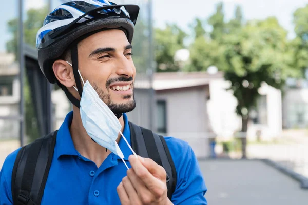 Веселый мусульманский курьер в велосипедном шлеме снимает медицинскую маску на открытом воздухе — стоковое фото