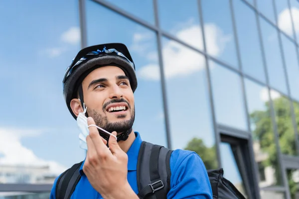 Sorridente fattorino arabo in casco da bici che toglie maschera protettiva all'aperto — Foto stock