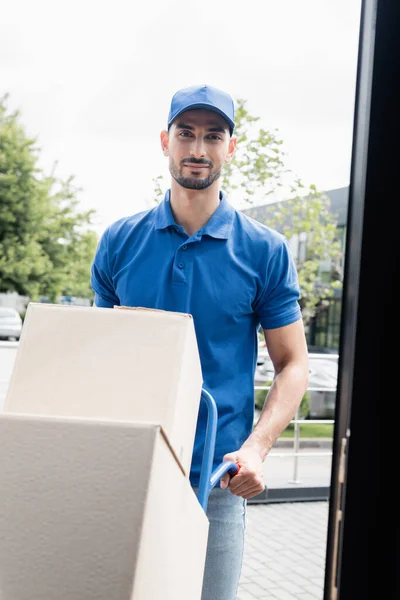 Arabian deliveryman looking at camera near cart with boxes — Stock Photo