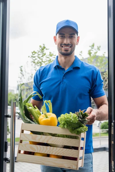 Livreur arabe souriant tenant boîte avec légumes près de la porte — Photo de stock