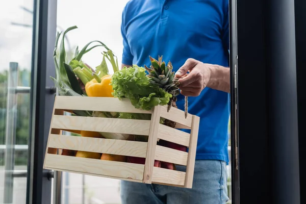 Обрезанный вид курьера, держащего коробку со свежими овощами у двери — стоковое фото
