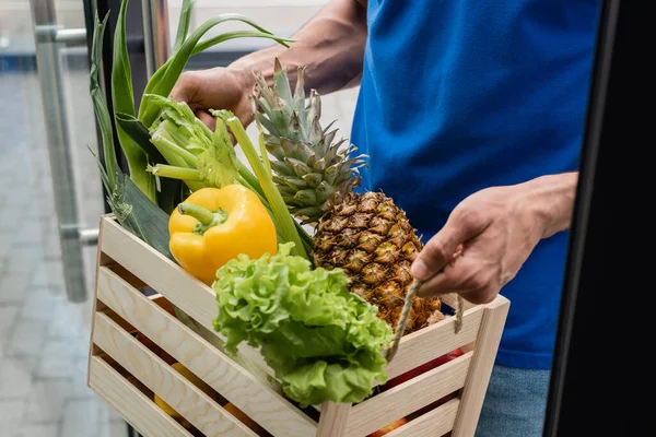 Обрезанный вид курьера, держащего коробку с органическими овощами и ананасом возле двери — стоковое фото
