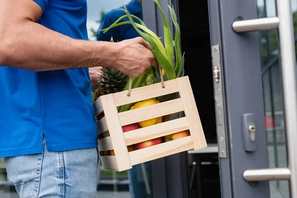 Ausgeschnittene Ansicht des Lieferanten hält Box mit frischen Lebensmitteln in der Nähe verschwommene Tür — Stockfoto