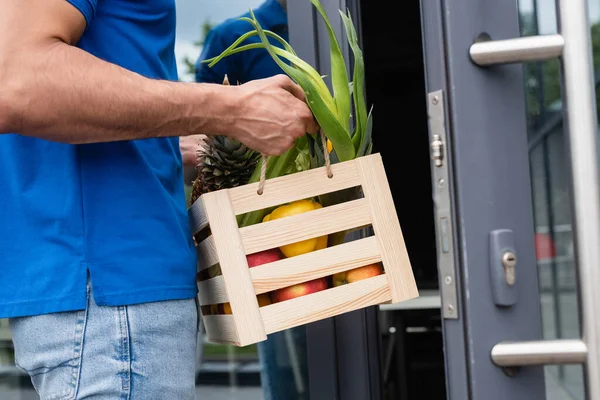 Vue recadrée du livreur tenant la boîte avec des aliments frais près de la porte floue — Photo de stock