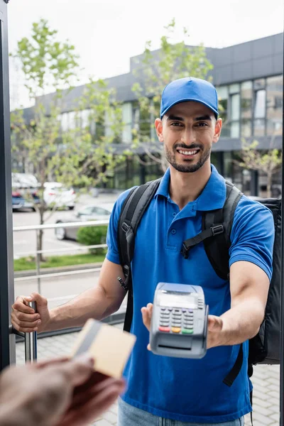 Sorrindo muçulmano entregador segurando terminal perto do cliente com cartão de crédito — Fotografia de Stock