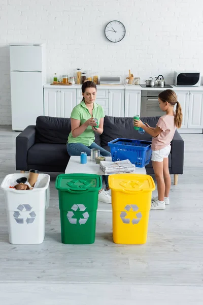 Woman and child holding tin cans near newspapers and trash bins with recycle sign — Stock Photo