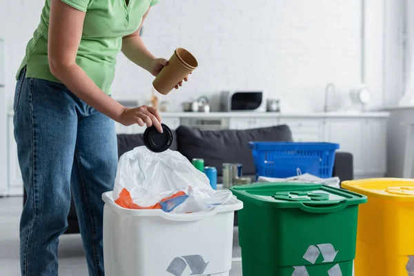 Обрезанный вид женщины, держащей мусор возле банки с табличкой переработки на кухне — стоковое фото