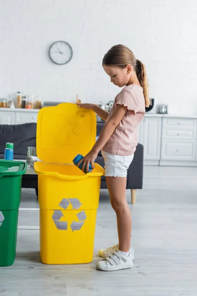 Vista lateral da criança colocando lata na lixeira com sinal de reciclagem em casa — Fotografia de Stock