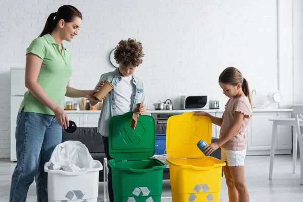 Femme souriante debout près des enfants avec poubelles et poubelles avec emblème de recyclage à la maison — Photo de stock