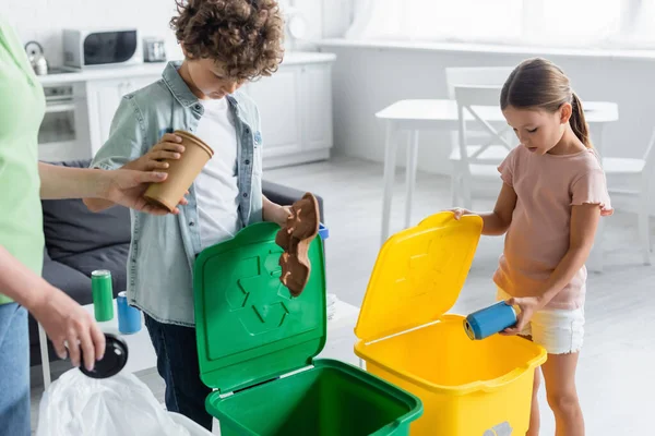 Bambini che selezionano rifiuti vicino alla madre e bidoni della spazzatura con segno di riciclaggio a casa — Foto stock