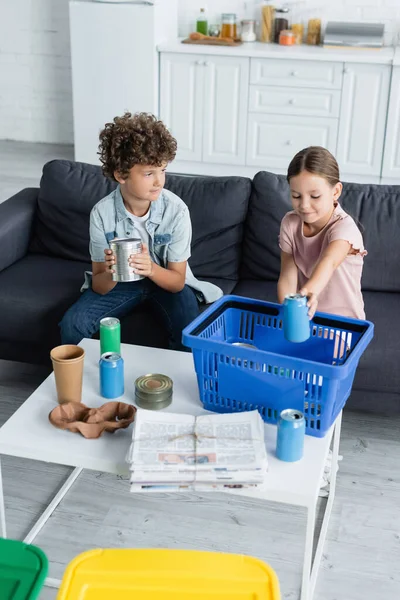 Chica poniendo lata en la cesta cerca de hermano y basura en casa - foto de stock