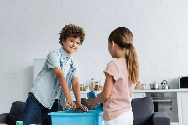 Garçon souriant mettre des boîtes de conserve dans la boîte près de soeur floue à la maison — Photo de stock