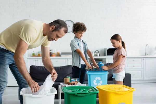 Hombre clasificando basura cerca de niños borrosos y botes de basura con letrero de reciclaje en casa - foto de stock