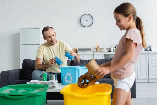 Sorrindo homem segurando latas de lata perto da caixa com sinal de reciclagem e filha borrada em casa — Fotografia de Stock