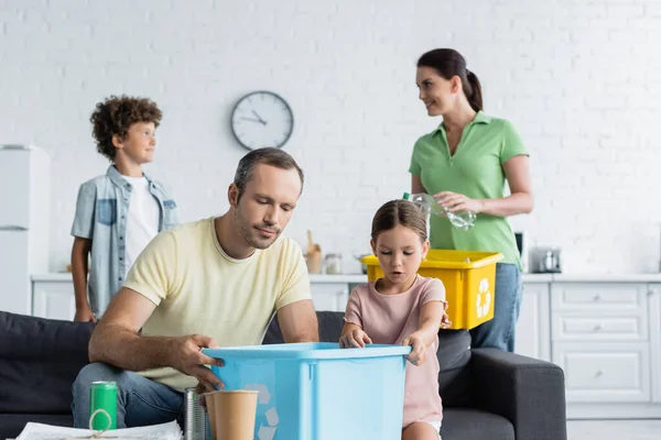 Père et fille tenant boîte avec panneau de recyclage près de la famille floue dans la cuisine — Photo de stock