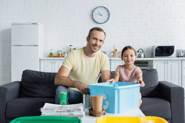 Alegre pai e filha segurando caixa com sinal de reciclagem perto de lixo na mesa na cozinha — Fotografia de Stock