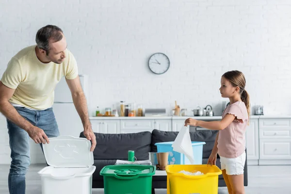 Padre e hija clasificando basura en botes de basura con letrero de reciclaje en la cocina - foto de stock
