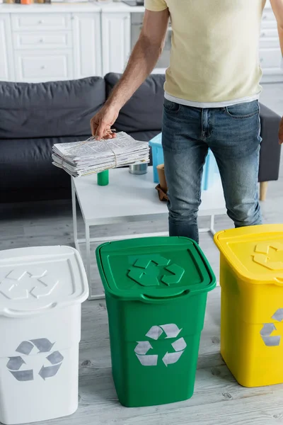 Vista cortada do homem segurando jornais perto de latas de lixo com sinal de reciclagem na cozinha — Fotografia de Stock