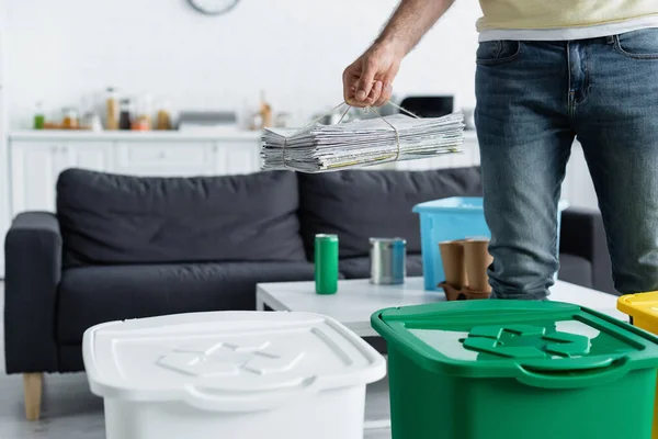 Cropped view of man holding newspapers near trash cans with recycle sign at home — Stock Photo