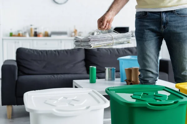 Vista recortada del hombre sosteniendo periódicos cerca de botes de basura con cartel de reciclaje en casa - foto de stock