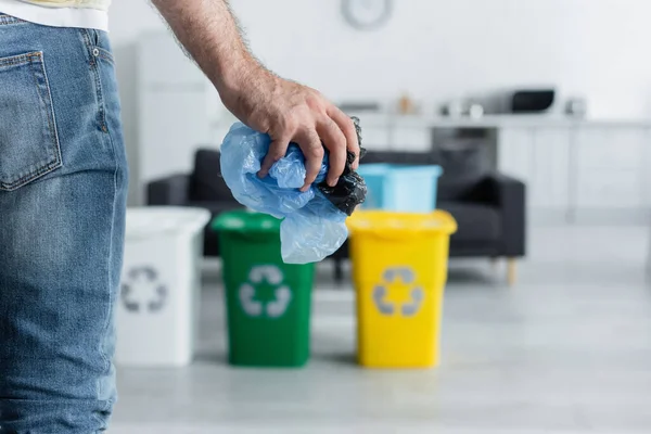Vista cortada do homem segurando sacos de plástico perto de latas borradas com sinal de reciclagem em casa — Fotografia de Stock