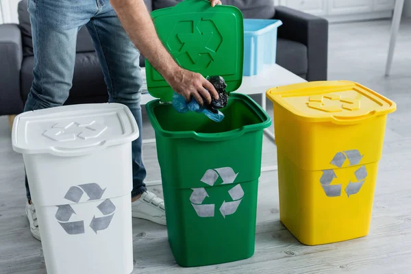 Vista recortada del hombre poniendo bolsas de plástico en la papelera con cartel de reciclaje en casa - foto de stock