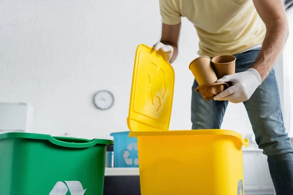 Vista recortada del hombre en guantes de látex sosteniendo vasos de papel cerca del bote de basura con cartel de reciclaje en casa - foto de stock