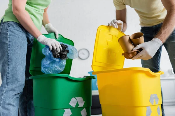 Vista cortada de casal em luvas de látex triagem de lixo em latas com sinal de reciclagem em casa — Fotografia de Stock