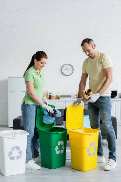 Lächelnde Frau mit Plastiktüten neben Ehemann in Latexhandschuhen und Mülltonnen mit Recyclingschild zu Hause — Stockfoto