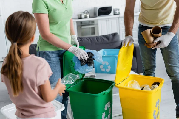 Батьки в латексних рукавичках сортують сміття біля розмитої дочки з пляшкою вдома — стокове фото