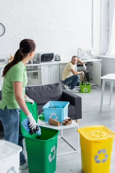 Homme en gants de latex regardant femme floue près des poubelles avec panneau de recyclage à la maison — Photo de stock