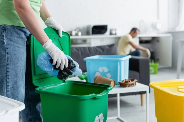 Cropped view of woman in latex gloves putting plastic bags in trash can with recycle sign — Stock Photo