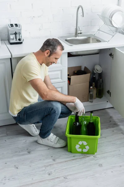 Vue latérale de l'homme souriant en gants de latex près des bouteilles en boîte avec panneau de recyclage — Photo de stock