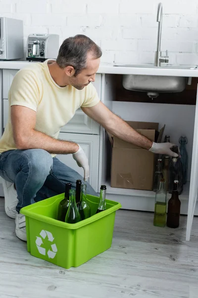 Homme prenant bouteille près de la boîte avec panneau de recyclage dans la cuisine — Photo de stock