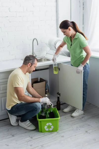 Vista lateral de la pareja sosteniendo botellas cerca de la caja con cartel de reciclaje en la cocina - foto de stock