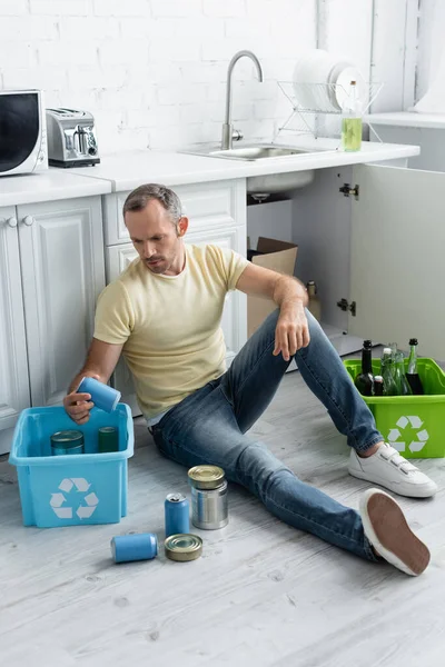 Homem sentado perto de caixas com sinal de reciclagem e lixo no chão na cozinha — Fotografia de Stock