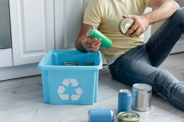 Vue recadrée de l'homme tenant des boîtes de conserve près de la boîte avec panneau de recyclage dans la cuisine — Photo de stock