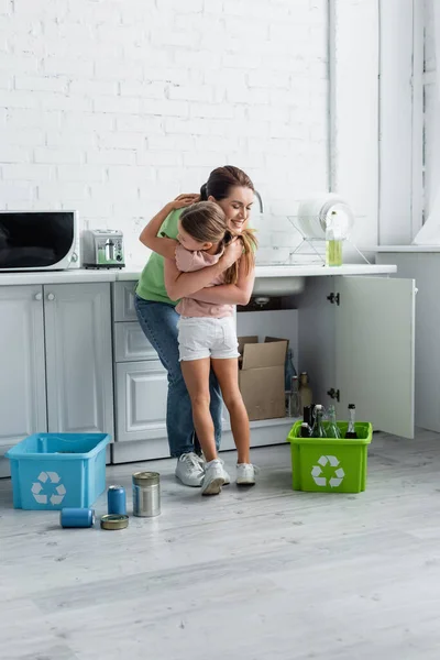 Mujer sonriente abrazando a su hija cerca de cajas con cartel de reciclaje en la cocina - foto de stock