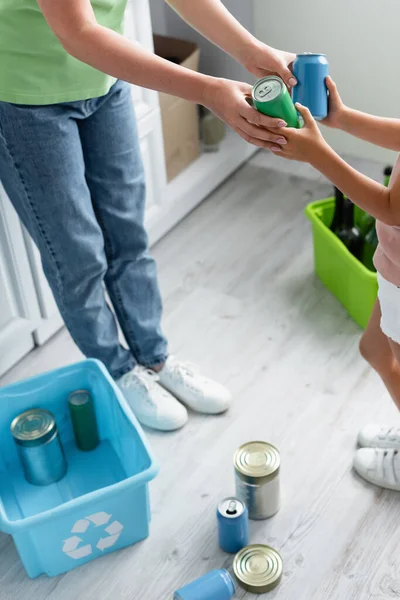 Vue recadrée de la femme et de la fille tenant des boîtes de conserve près des boîtes avec panneau de recyclage dans la cuisine — Photo de stock