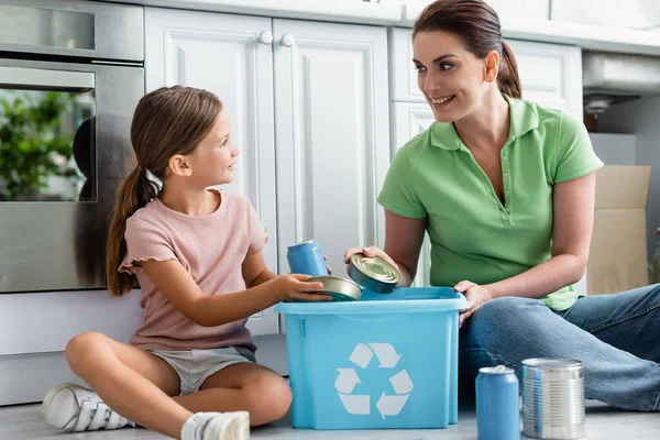 Smiling woman and daughter holding tin cans near box with recycle sign in kitchen — Stock Photo