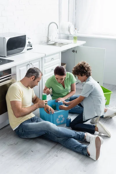 Familia con hijo poniendo latas de lata en caja con cartel de reciclaje en la cocina - foto de stock