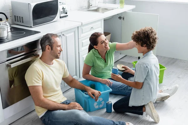 Donna sorridente che tocca il figlio con lattine vicino al marito e scatole con simbolo del riciclo in cucina — Foto stock