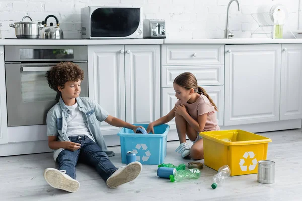 Niños poniendo latas en caja con cartel de reciclaje en el suelo en la cocina - foto de stock
