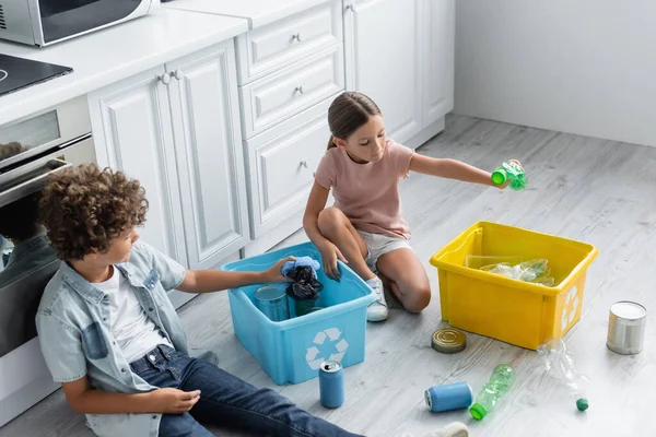 Vue grand angle de la fille tenant bouteille près du frère et des boîtes avec panneau de recyclage dans la cuisine — Photo de stock