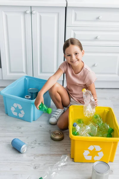 Smiling girl holding bottles near boxes with recycle sign in kitchen — Stock Photo