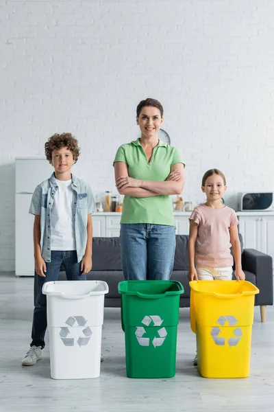 Donna sorridente guardando la fotocamera vicino ai bambini e bidoni della spazzatura con segno di riciclaggio in cucina — Foto stock