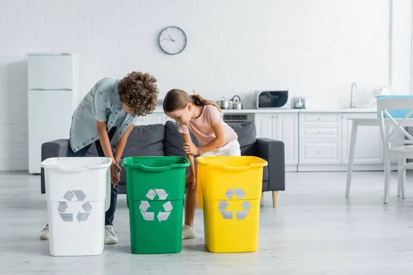 Niños mirando botes de basura con cartel de reciclaje en la cocina - foto de stock