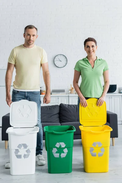 Mujer sonriente mirando a la cámara cerca de marido y botes de basura con cartel de reciclaje en la cocina - foto de stock