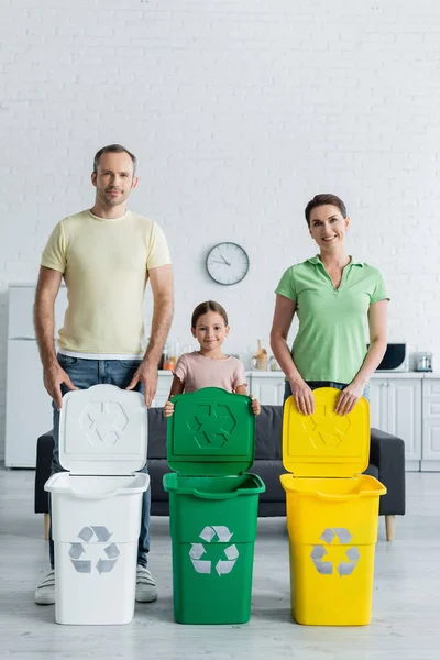 Famille positive avec fille debout près des poubelles avec panneau de recyclage dans la cuisine — Photo de stock